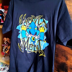 May include: Navy blue t-shirt featuring a graphic of a blue robot with yellow accents and the word "Metal" in a stylized font. The design is set against a blue and white background. The shirt is hanging on a white hanger.