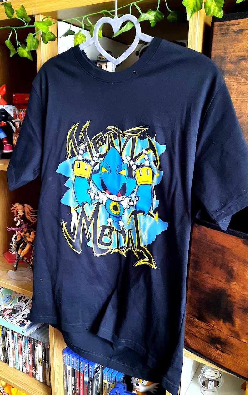 May include: Navy blue t-shirt featuring a graphic of a blue robot with yellow accents and the word "Metal" in a stylized font. The design is set against a blue and white background. The shirt is hanging on a white hanger.