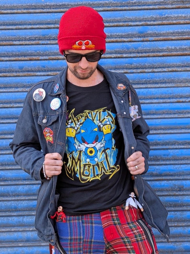 May include: A person wearing a red knit beanie, black sunglasses, and a black t-shirt with a blue and yellow graphic of a robot. The person is also wearing a denim jacket with patches and plaid pants.
