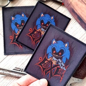 May include: Square patches featuring a blue cartoon character with red eyes and a sinister grin. The patches have a dark background with red accents and are surrounded by a black border. The patches are displayed near a silver revolver.