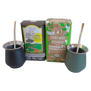 May include: Two mate gourds with metal bombillas, one black and one green, accompanied by two 500g packages of organic yerba mate. The packages are labeled La Oberena Organica and Amanda, featuring printed text, graphics of trees, and leaves.