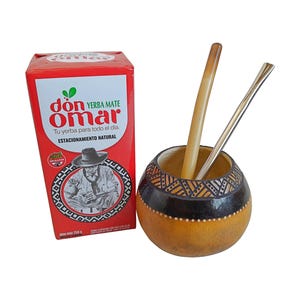 Può includere: Una scatola rossa di yerba mate Don Omar con etichetta da 250g e una zucca mate rotonda intagliata con un motivo geometrico. All'interno della zucca ci sono due cannucce, una in materiale chiaro e l'altra in metallo color argento.