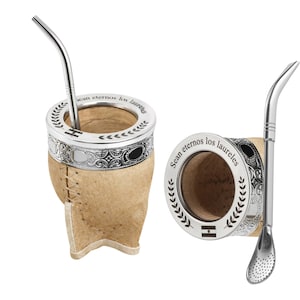 May include: Two yerba mate gourds with metal rims and straws. The gourds are made of natural tan leather and have a stitched seam. The metal rims have decorative patterns and the text "Sean eternos los laureles". One gourd has a straw inserted.