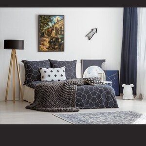 May include: A bedroom featuring a bed with dark gray bedding patterned with white geometric hexagons, a white throw pillow with black plus signs, and a chunky knit dark gray blanket. A framed painting of a street scene hangs on the wall.