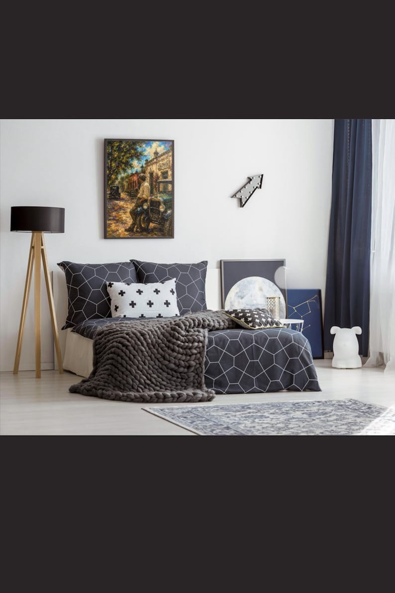 May include: A bedroom featuring a bed with dark gray bedding patterned with white geometric hexagons, a white throw pillow with black plus signs, and a chunky knit dark gray blanket. A framed painting of a street scene hangs on the wall.