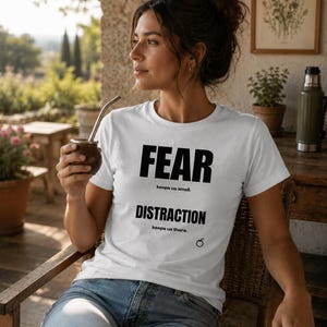 May include: A white short-sleeved t-shirt featuring black text that reads FEAR keeps us small. DISTRACTION keeps us there. The shirt includes a small black circular graphic near the bottom right corner of the printed text area.