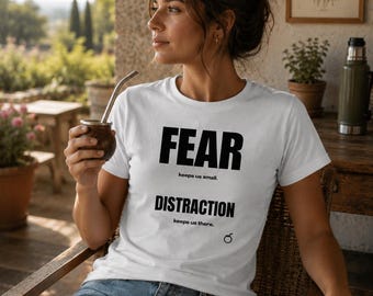 May include: A white short-sleeved t-shirt featuring black text that reads FEAR keeps us small. DISTRACTION keeps us there. The shirt includes a small black circular graphic near the bottom right corner of the printed text area.