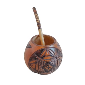 May include: A round, brown gourd mate cup featuring intricate dark brown carved geometric patterns on its exterior surface. A slender, light-colored wooden bombilla straw with a dark brown patterned tip is inserted into the open top of the cup.