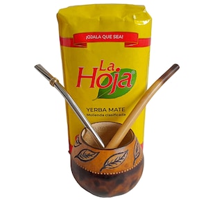 May include: A yellow La Hoja Yerba Mate package with red text and a green leaf logo, positioned behind a round, brown gourd with leaf carvings. Two metal bombilla straws, one silver-toned and one light brown, are inserted into the gourd opening.