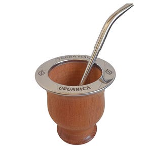 May include: A wooden yerba mate cup with a stainless steel rim and straw. The rim is engraved with the words "YERBA MAT" and "ORGANICA". The straw is curved and has a silver finish. The cup is made of light brown wood.