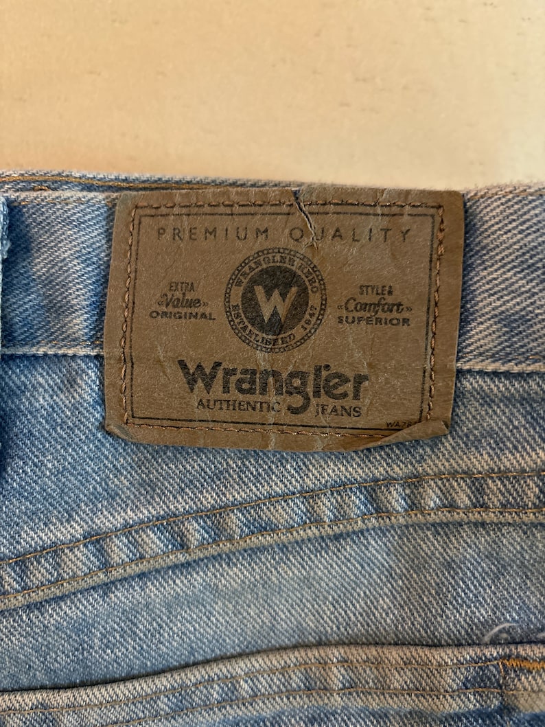 May include: A close-up of a brown leather patch sewn onto a pair of blue denim jeans. The patch has the text "Wrangler Authentic Jeans" and a logo with the text "Wrangler" and "Established 1947". The patch also has the text "Premium Quality", "Extra Value Original", and "Style & Comfort Superior".