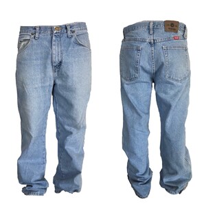 Puede incluir: Vaqueros de mezclilla azul claro, vista frontal y trasera, con un dise&ntilde;o cl&aacute;sico de cinco bolsillos. Los vaqueros tienen cierre de bot&oacute;n y cremallera, trabillas para el cintur&oacute;n y un parche de cuero marr&oacute;n con el logo de Wrangler en la parte trasera.