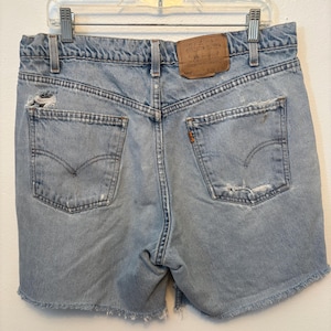 May include: Light blue denim shorts with a high-waisted fit, featuring a brown leather patch on the back waistband, two back pockets with stitched arcuate designs, frayed raw hems, and distressed areas on the pockets and upper left side.