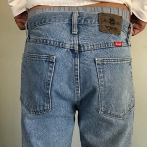 Puede incluir: Vaqueros de mezclilla azul claro con dos bolsillos traseros y una etiqueta Wrangler de cuero marr&oacute;n. Los vaqueros tienen un corte cl&aacute;sico y un lavado descolorido. La etiqueta Wrangler es visible en la cintura trasera.