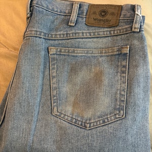 May include: A pair of light blue denim jeans with a brown leather patch on the back pocket that says "Wrangler Authentic Jeans" in white text.