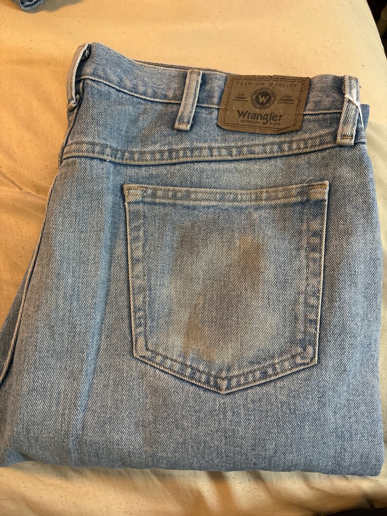 May include: A pair of light blue denim jeans with a brown leather patch on the back pocket that says "Wrangler Authentic Jeans" in white text.