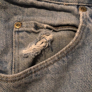 Puede incluir: Primer plano de un bolsillo de vaqueros azul claro con un borde desgastado y deshilachado. El bolsillo tiene un peque&ntilde;o agujero y est&aacute; cosido con un hilo en contraste. Los vaqueros tienen un dise&ntilde;o cl&aacute;sico de cinco bolsillos.