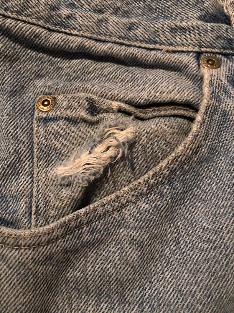 Puede incluir: Primer plano de un bolsillo de vaqueros azul claro con un borde desgastado y deshilachado. El bolsillo tiene un peque&ntilde;o agujero y est&aacute; cosido con un hilo en contraste. Los vaqueros tienen un dise&ntilde;o cl&aacute;sico de cinco bolsillos.