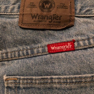 Puede incluir: Primer plano de vaqueros de mezclilla azul claro. Se ve un parche de cuero marr&oacute;n con el logotipo de Wrangler, junto con una etiqueta roja con el logotipo de Wrangler. Los vaqueros tienen un aspecto cl&aacute;sico y desgastado.