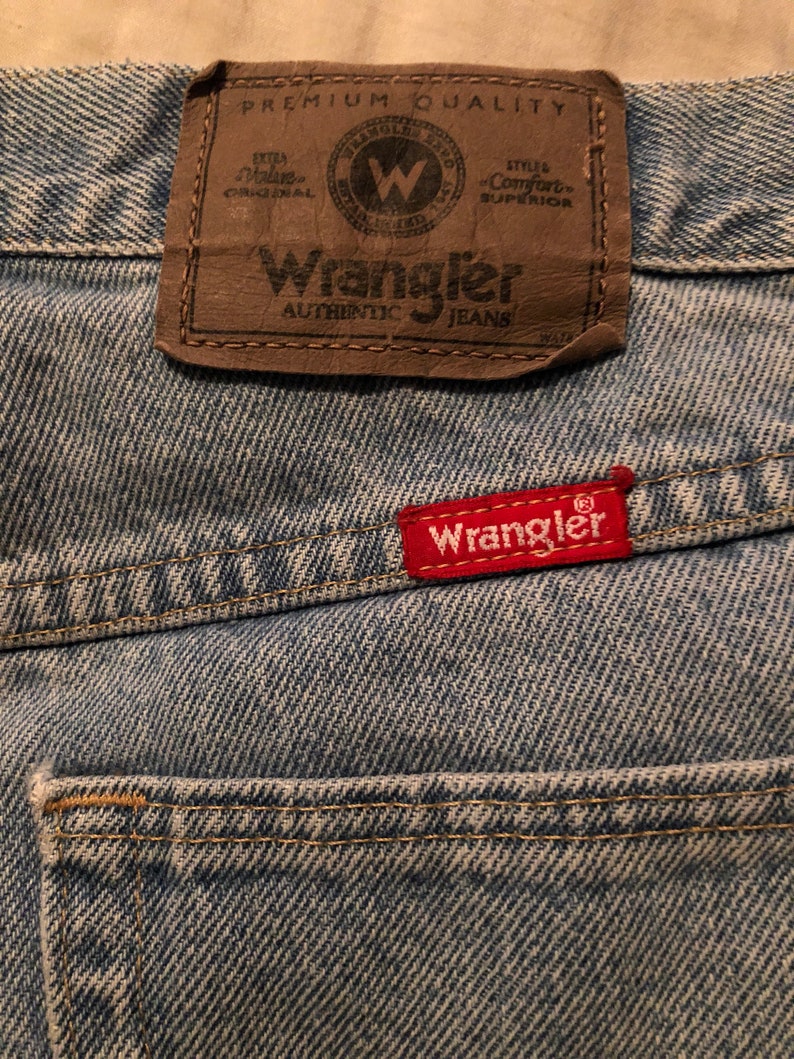 Puede incluir: Primer plano de vaqueros de mezclilla azul claro. Se ve un parche de cuero marr&oacute;n con el logotipo de Wrangler, junto con una etiqueta roja con el logotipo de Wrangler. Los vaqueros tienen un aspecto cl&aacute;sico y desgastado.