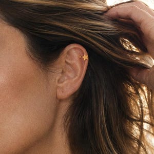 May include: A small gold-toned ear cuff featuring a five-pointed star design. The jewelry piece is worn on the upper cartilage of the ear, showcasing a metallic finish and a simple, clean shape that rests securely against the outer ear edge.