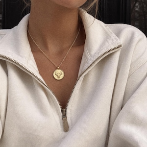 May include: A gold-toned necklace featuring a circular pendant with an embossed bull design, worn with a cream-coloured, long-sleeved, quarter-zip sweatshirt that has a silver-toned metal zip pull and a high collar neckline.