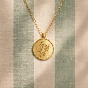Puede incluir: Collar con colgante dorado con un dije redondo con un diseño detallado de escorpión. El colgante cuelga de una delicada cadena dorada. El fondo es un patrón de rayas en blanco y azul claro.