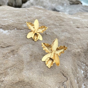 Puede incluir: Dos pendientes dorados con forma de flor. Los pendientes presentan pétalos texturizados y un estambre central. Se exhiben sobre una superficie de piedra texturizada de color marrón claro, con el mar al fondo.