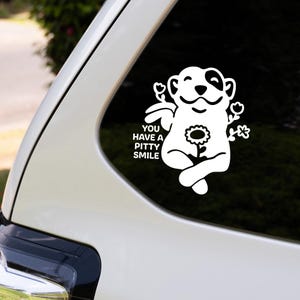 May include: A white vinyl decal applied to a car window featuring a cartoon dog with a patch over one eye, sitting with crossed legs, holding a flower, and surrounded by small floral accents with the text YOU HAVE A PITTY SMILE printed clearly.