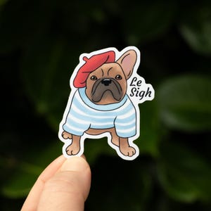 May include: A sticker featuring a brown French Bulldog wearing a red beret and a blue and white striped shirt. The sticker has the text "Le Sigh" in a white font.