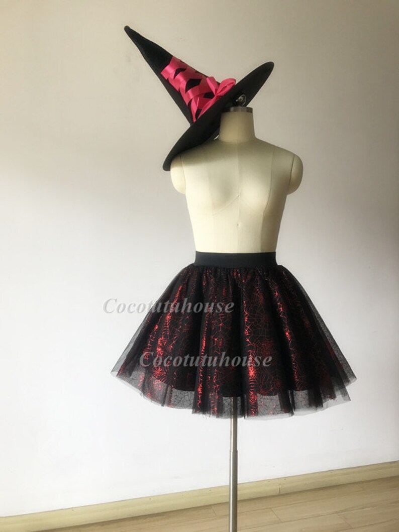 Pu&ograve; includere: Gonna di tulle nera con un motivo a ragnatela rosso. La gonna &egrave; su un manichino e ha una cintura nera. Un cappello da strega nero con un nastro rosa &egrave; sulla testa del manichino.
