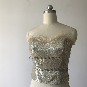 May include: A gold sequined strapless top with a nude tulle ruffle trim. The top is on a white mannequin.
