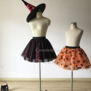 Pu&ograve; includere: Due gonne di tulle con stampe a tema Halloween. La gonna a sinistra &egrave; nera con ragnatele rosse e la gonna a destra &egrave; arancione con pipistrelli neri, zucche e ragni. Entrambe le gonne sono su manichini con supporti neri.