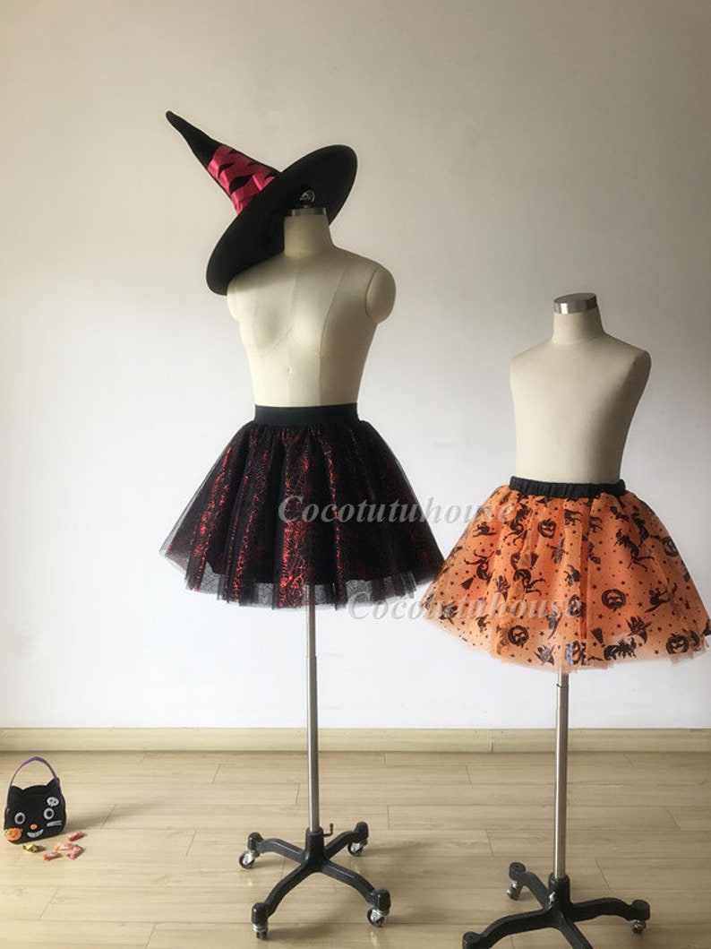 Pu&ograve; includere: Due gonne di tulle con stampe a tema Halloween. La gonna a sinistra &egrave; nera con ragnatele rosse e la gonna a destra &egrave; arancione con pipistrelli neri, zucche e ragni. Entrambe le gonne sono su manichini con supporti neri.