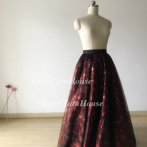 Pu&ograve; includere: Una lunga gonna di tulle nera con un motivo a ragnatela rosso. La gonna ha una cintura nera ed &egrave; mostrata su un manichino bianco.