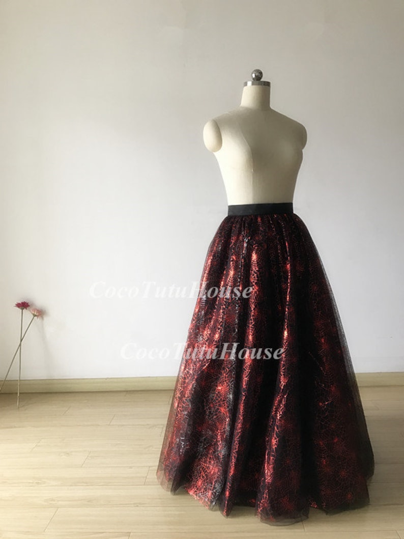 Pu&ograve; includere: Una lunga gonna di tulle nera con un motivo a ragnatela rosso. La gonna ha una cintura nera ed &egrave; mostrata su un manichino bianco.