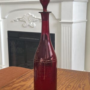 May include: A tall, ruby-red glass decanter with a matching stopper. The decanter features a slender neck and a bulbous base, with gold-colored floral detailing. The stopper is a teardrop shape. The decanter is approximately 12 inches tall.
