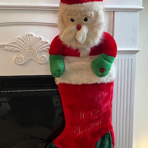 May include: A plush Christmas stocking shaped like Santa Claus. The stocking is red with green accents and features the embroidered words "Merry Christmas". Santa has a red hat, white beard, and green mittens.