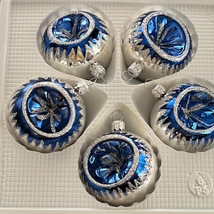 May include: Five glass Christmas ornaments in a white tray. Each ornament is silver with a blue center and silver glitter accents. The ornaments have a classic round shape and are ready to hang.