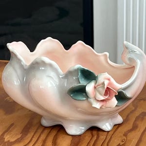 Könnte beinhalten: Dekorative Keramikschale in zarten Rosa- und Grautönen, mit einer geformten Rose und grünen Blättern. Die Schale hat einen gewellten Rand und steht auf drei kleinen Füßen, für die Inneneinrichtung.