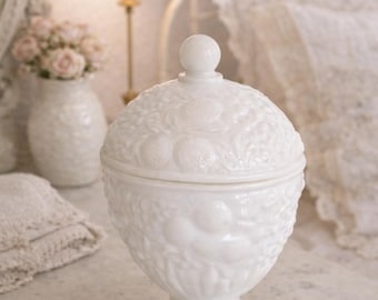 May include: A white, lidded jar with a pedestal base. The jar features a detailed, embossed pattern of fruit and foliage. The lid is topped with a small, round knob. The jar is on a white surface.