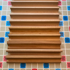 May include: A wooden Scrabble tile rack with a curved design, resting on a Scrabble game board. The rack has multiple grooves to hold game tiles. The board features the Scrabble logo and game board squares with point multipliers.