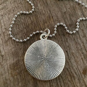 Peut inclure: Collier avec pendentif argent&eacute; et pendentif rond textur&eacute;. Le pendentif pr&eacute;sente un motif radial et est suspendu &agrave; une fine cha&icirc;ne &agrave; billes. Le collier est pr&eacute;sent&eacute; sur une surface en bois.