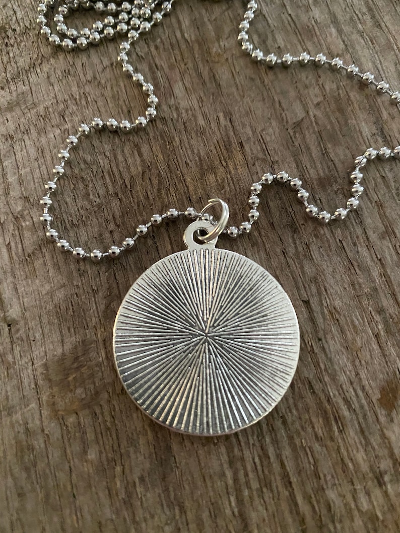 Peut inclure: Collier avec pendentif argent&eacute; et pendentif rond textur&eacute;. Le pendentif pr&eacute;sente un motif radial et est suspendu &agrave; une fine cha&icirc;ne &agrave; billes. Le collier est pr&eacute;sent&eacute; sur une surface en bois.
