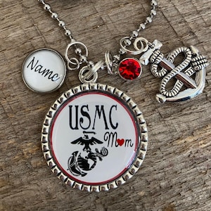 May include: A silver-toned pendant necklace featuring a round charm with "USMC Mom" text and a Marine Corps emblem. Includes a small charm with the word "Name", a red gem, and an anchor charm. The background is a wooden surface.