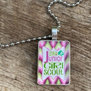 Pode incluir: Um colar de corrente prateada com um pingente retangular com um design estampado em rosa, verde e branco. O pingente tem o texto "I'm a Junior Girl Scout" com um logotipo verde de Girl Scout.