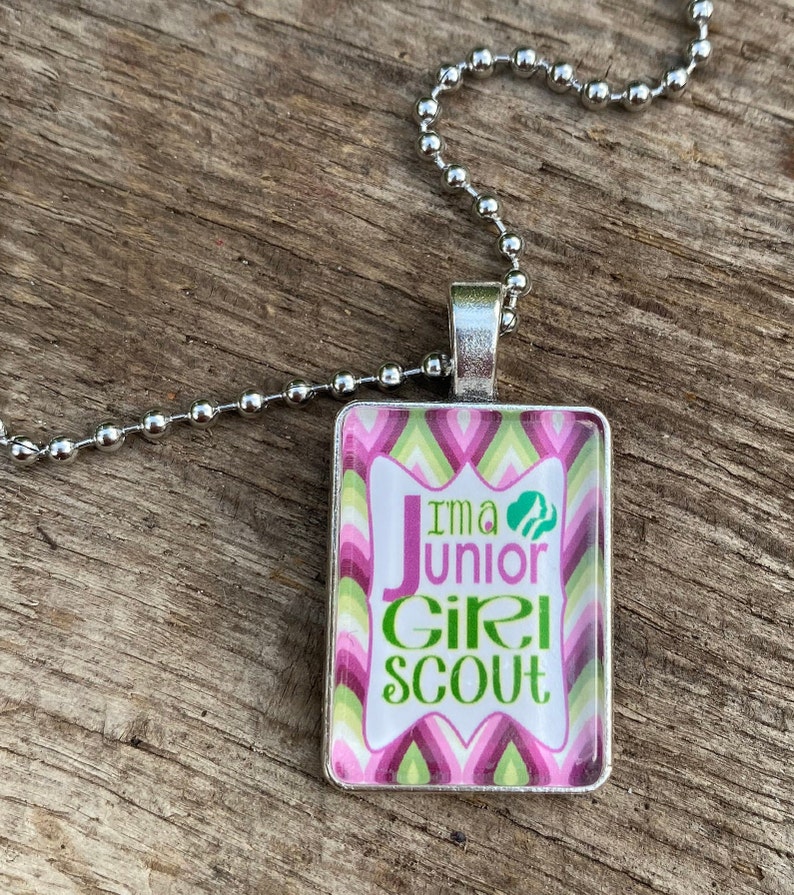 Pode incluir: Um colar de corrente prateada com um pingente retangular com um design estampado em rosa, verde e branco. O pingente tem o texto "I'm a Junior Girl Scout" com um logotipo verde de Girl Scout.