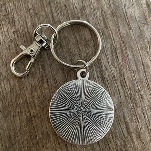 Peut inclure: Porte-cl&eacute;s argent&eacute; avec un pendentif rond &agrave; motif radial. Le porte-cl&eacute;s comprend un anneau bris&eacute; et un mousqueton pour une fixation facile. Le design du pendentif a un aspect textur&eacute; et vintage.
