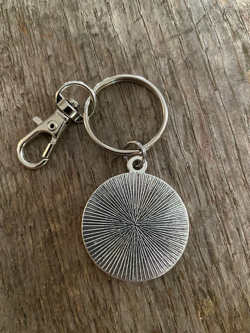 Peut inclure: Porte-cl&eacute;s argent&eacute; avec un pendentif rond &agrave; motif radial. Le porte-cl&eacute;s comprend un anneau bris&eacute; et un mousqueton pour une fixation facile. Le design du pendentif a un aspect textur&eacute; et vintage.