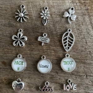 Peut inclure: Une collection de breloques argent&eacute;es de diff&eacute;rentes formes, dont des fleurs, un tr&egrave;fle, une feuille et des c&oelig;urs. Certaines breloques portent les mots "Pack", "Name", "Troop" et "Love". Les breloques sont pr&eacute;sent&eacute;es sur une surface en bois.